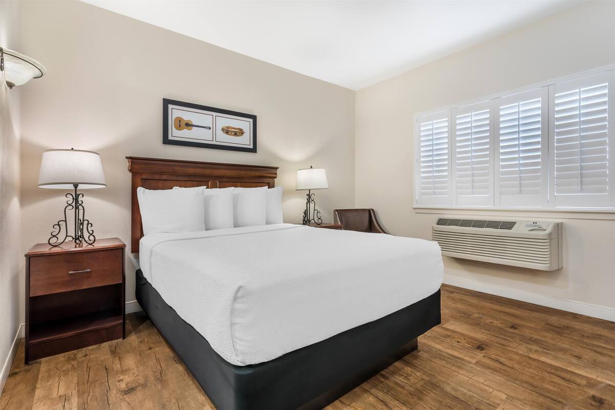 A hotel room with a large bed, two bedside tables with lamps, a framed picture above the bed, a window with blinds, and a wall-mounted air conditioner.