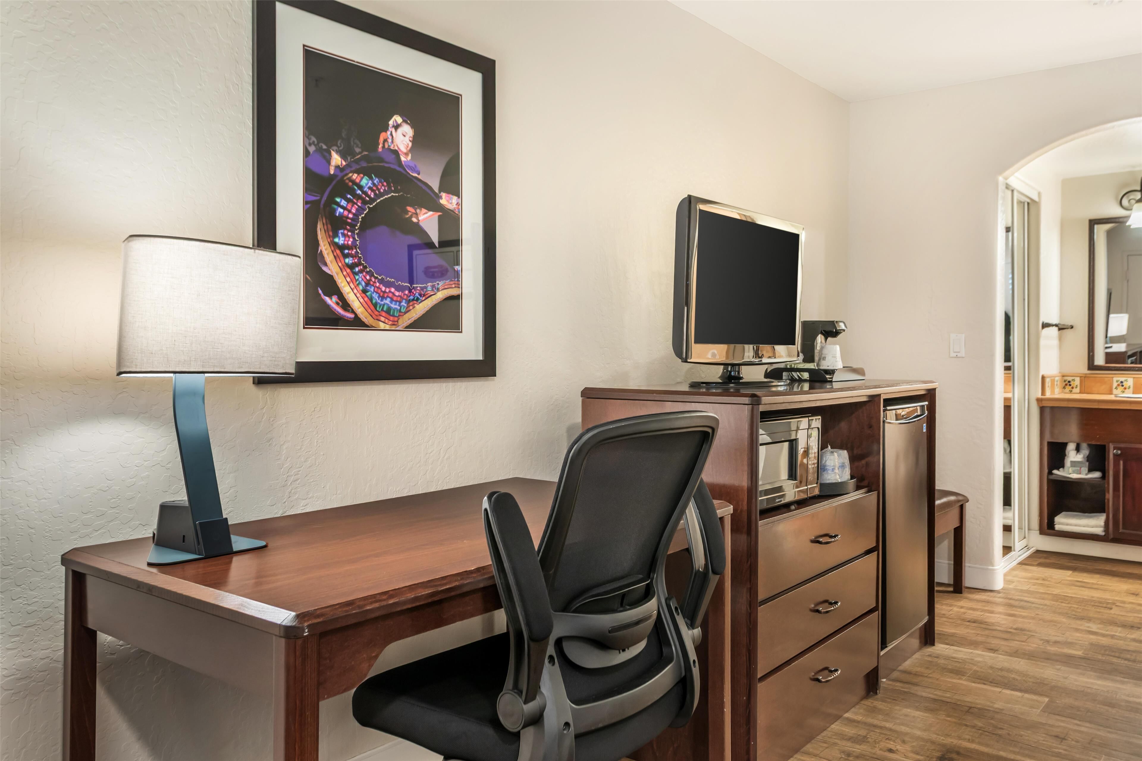 Ein Hotelzimmer mit einem Holztisch, einem schwarzen Bürostuhl, einem gerahmten Bild an der Wand, einem Flachbildfernseher, einer Kaffeemaschine und einem Holzschrank.