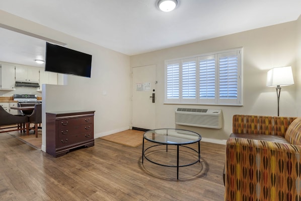 A living room with a sofa, a glass coffee table, a TV, a kitchen area, and a door with a window.