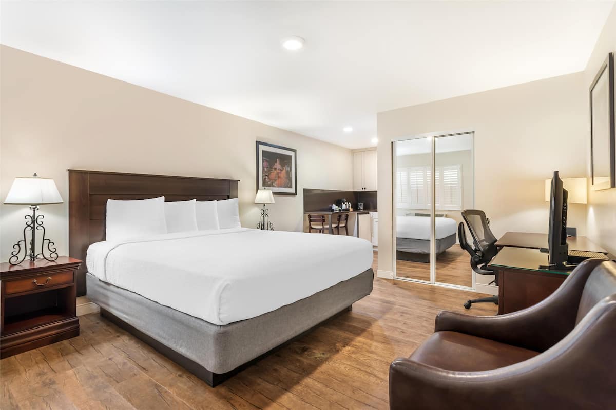 A hotel room with a large bed, a desk with a computer, a chair, a nightstand, a lamp, and a mirror.