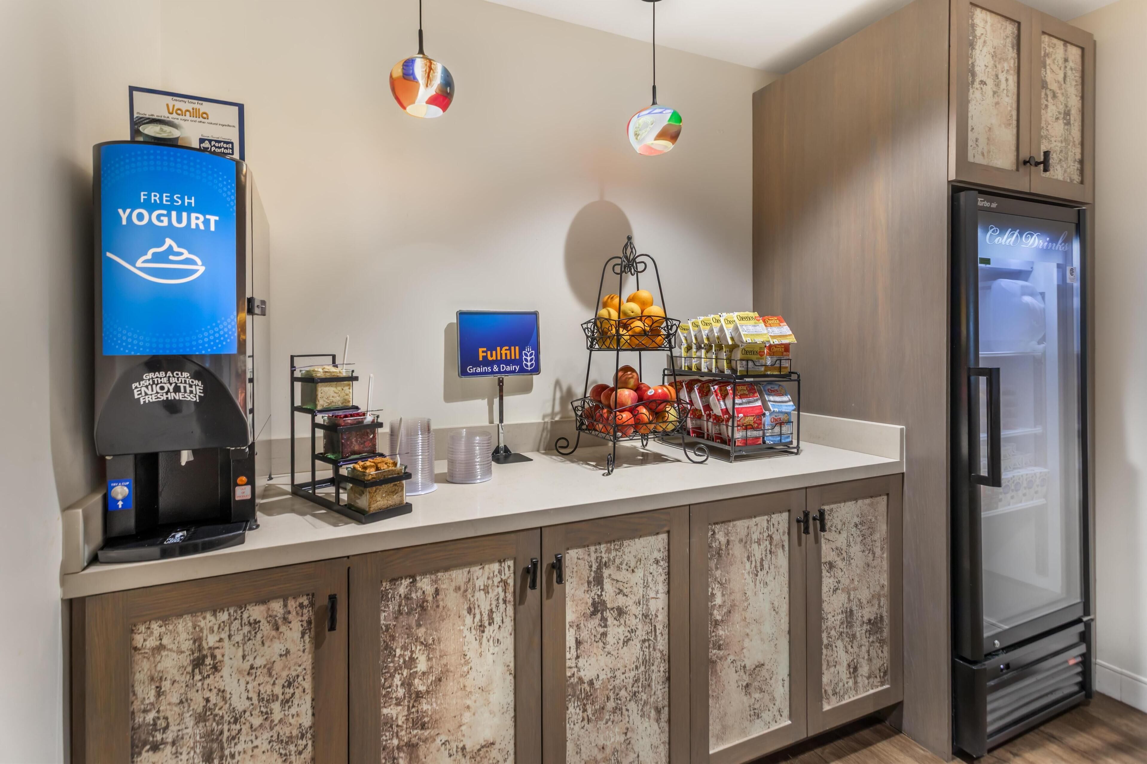 A self-service breakfast area with a yogurt dispenser, a coffee machine, a fruit display, and a refrigerated drink cooler.