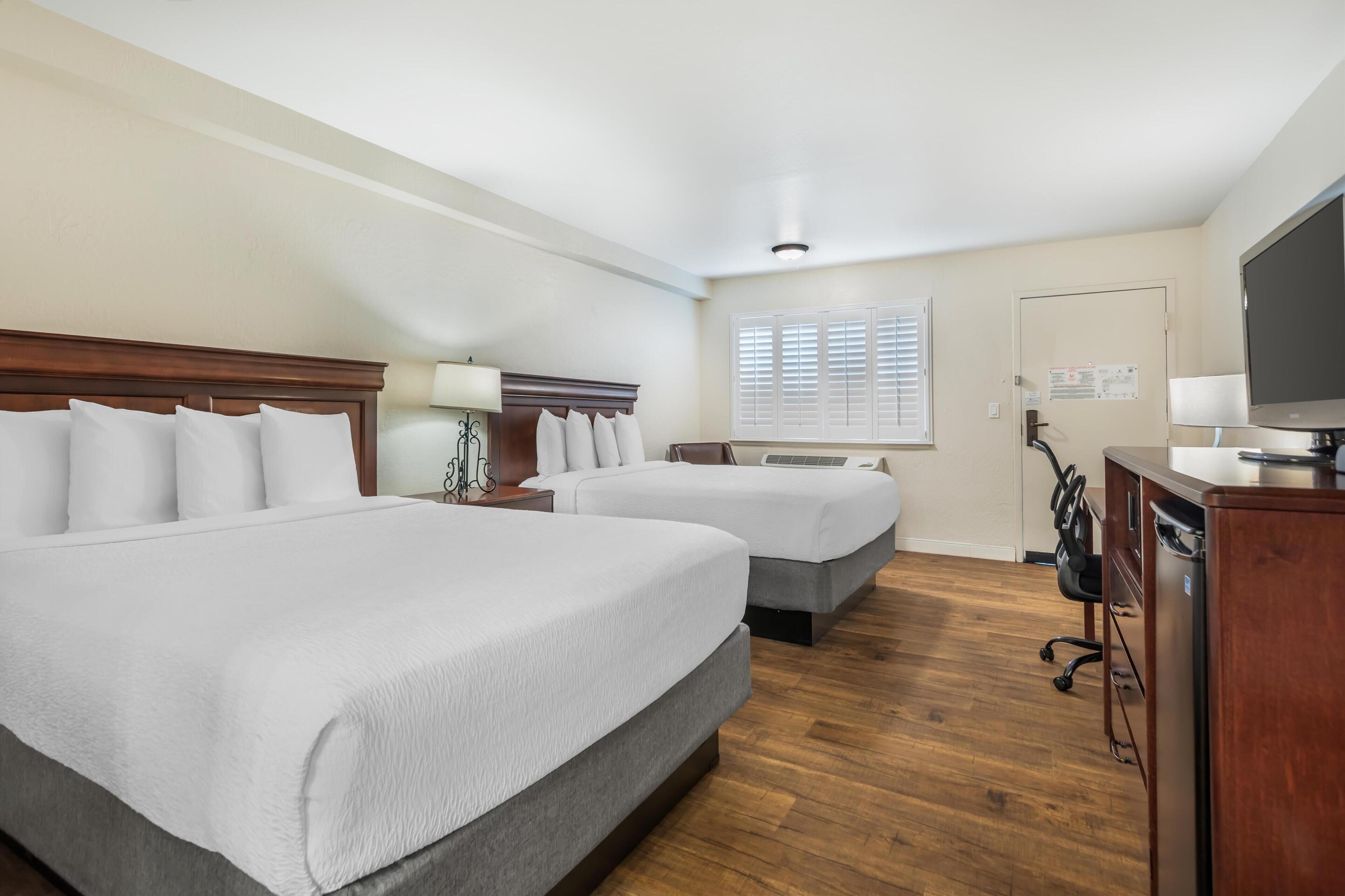 Quarto de hotel com duas camas, uma mesa de escritório com computador, televisão e janela com persianas.