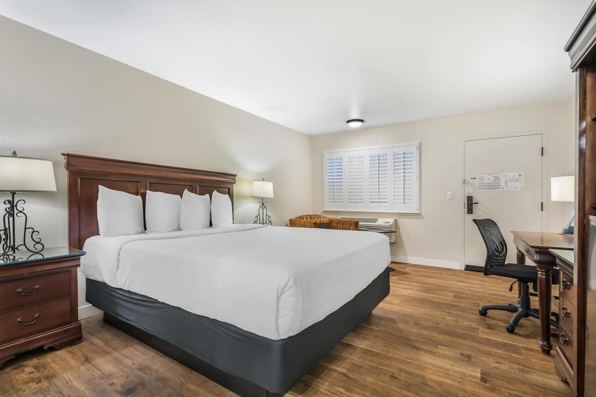 A bedroom with a large bed, bedside tables with lamps, a desk with a chair, and a window with blinds.