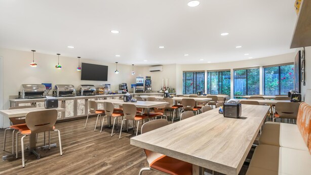 A modern dining area with multiple tables and chairs, a kitchen area with appliances, and large windows providing natural light. - Best Western Rose Garden Inn (Watsonville)