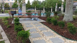 Un sendero pavimentado con piedras, rodeado de acolchado y arbustos pequeños, que lleva a una mesa de picnic y una zona de piscina, todo ello delimitado por una valla de metal negro.