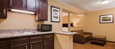 A compact kitchen with dark cabinets, a microwave, and a sink. There is a framed picture on the wall and a small table with a lamp in the living area.