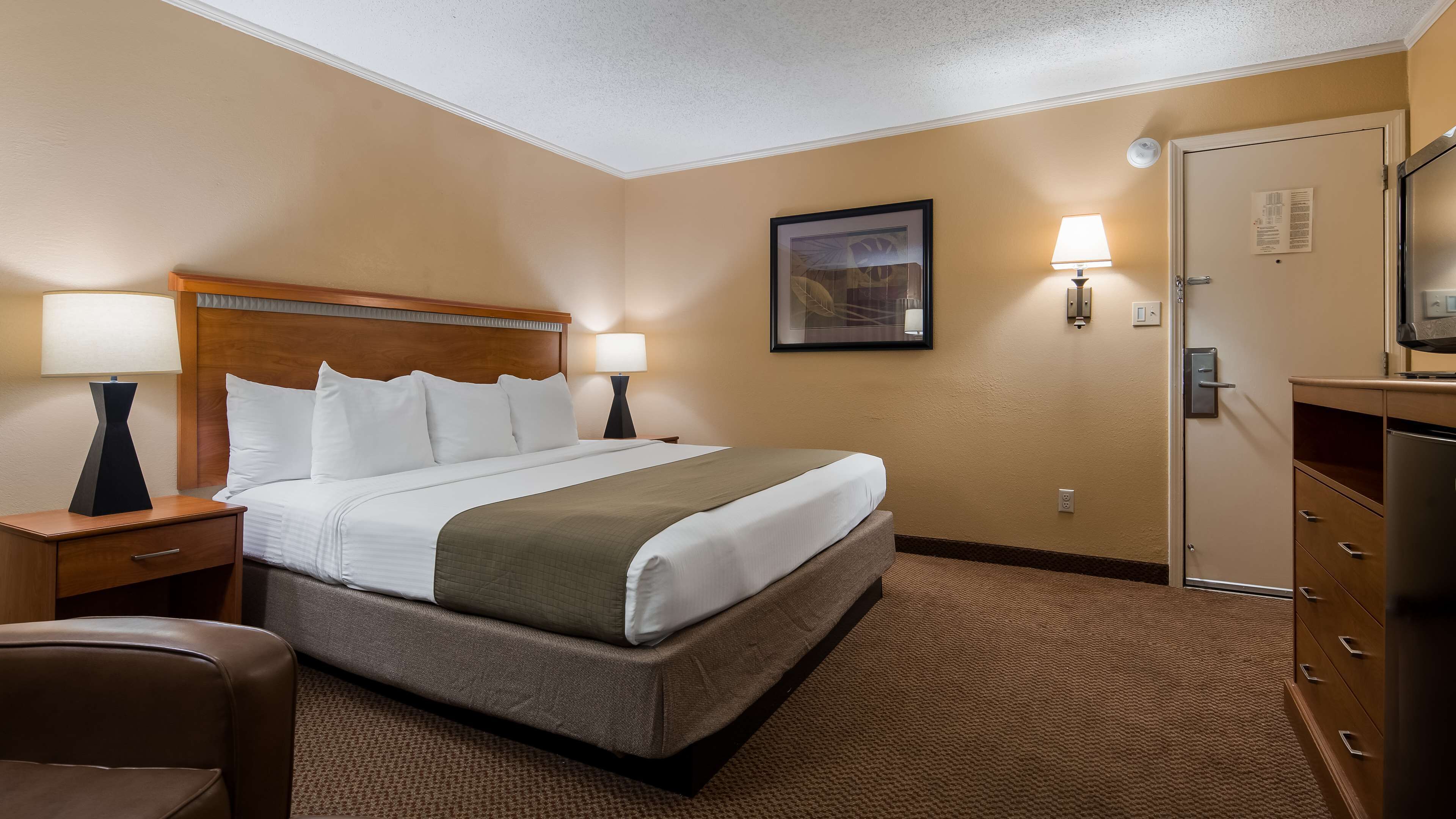 A hotel room with a large bed, two bedside tables with lamps, a wooden headboard, a framed picture on the wall, a door with a mirror, and a small desk with drawers.