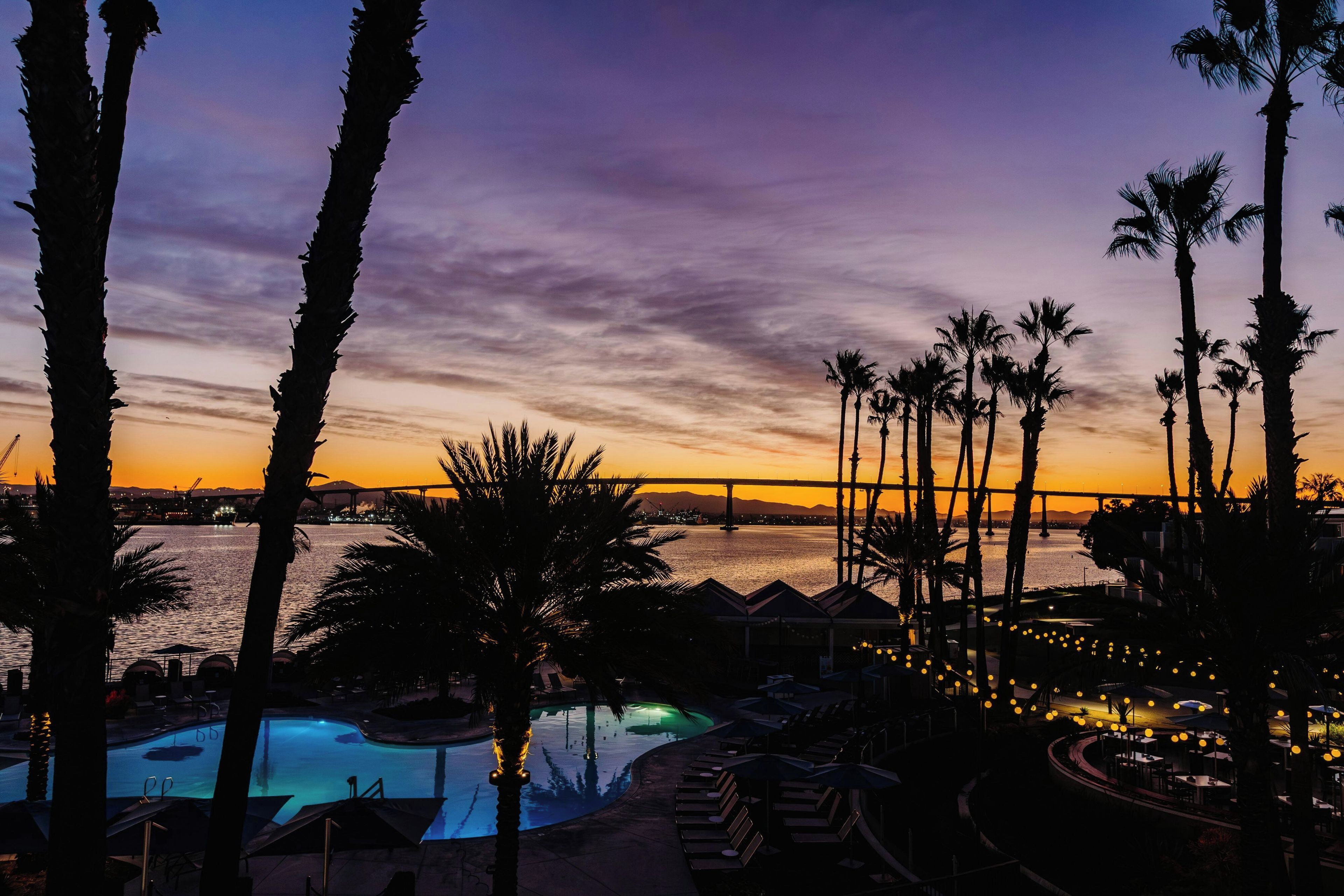 A resort pool area with lounge chairs and umbrellas, overlooking a body of water and a bridge.
