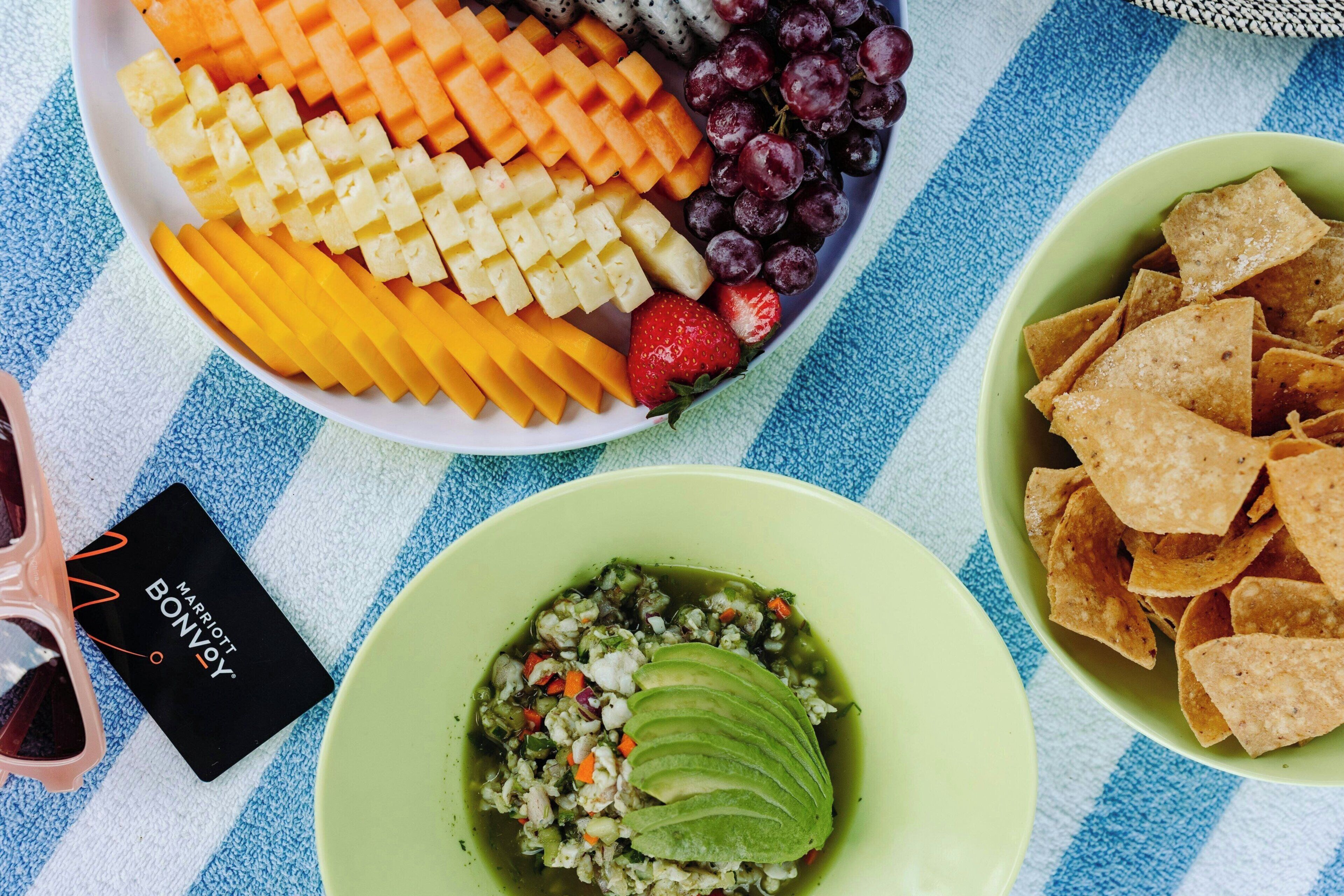 Une table sur laquelle on trouve une variété d’aliments, notamment des fruits, des chips et un plat vert contenant une tranche d’avocat.