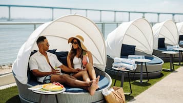 A couple relaxing in a covered outdoor seating area with a view of a bridge, surrounded by lounge chairs and tables.