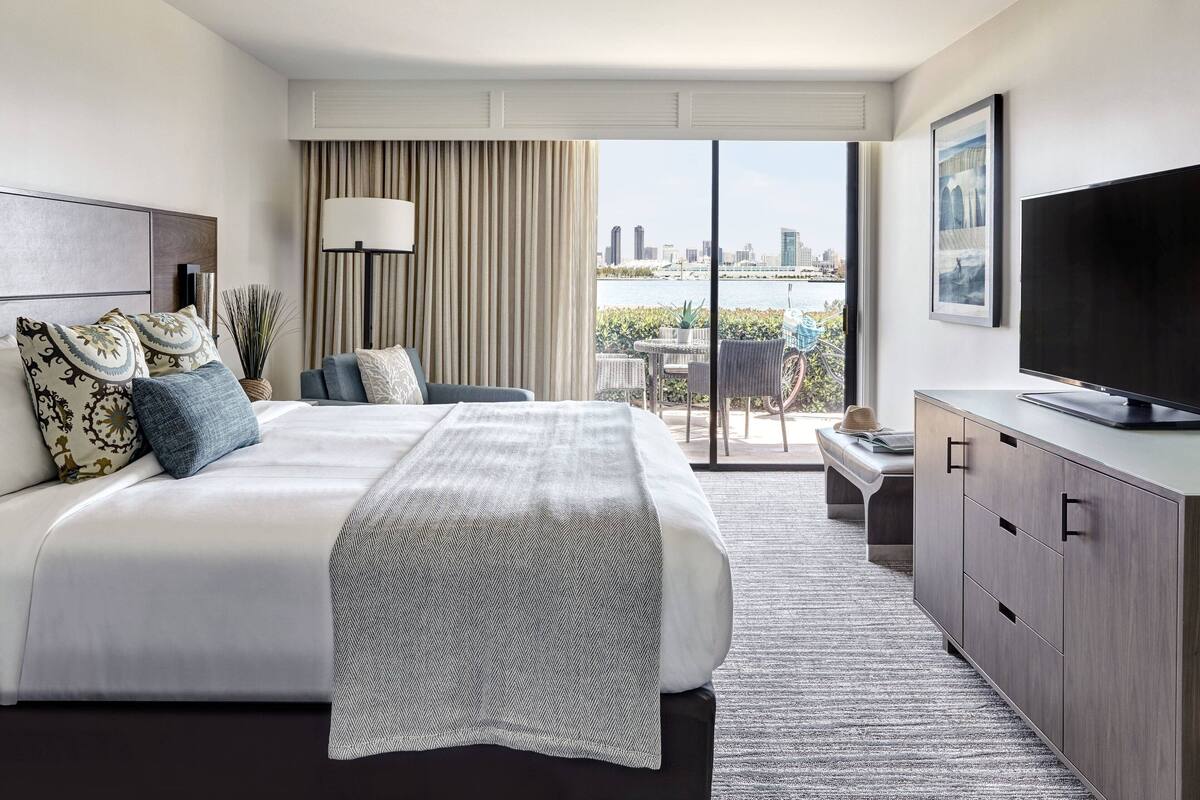 A hotel room with a large bed, a TV, a balcony with a view of the city, and a desk with a chair.