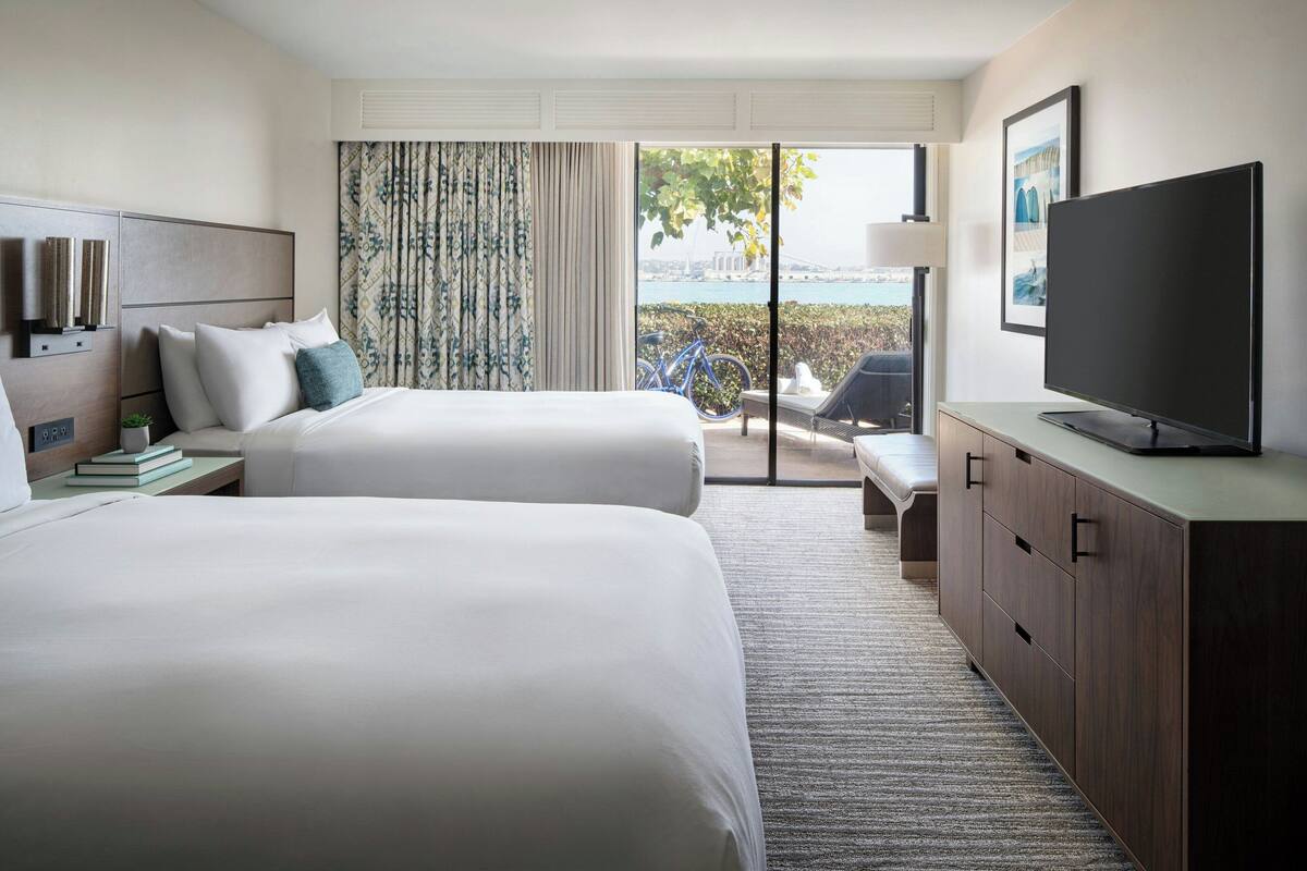 A hotel room with two beds, a TV, a desk, and a view of the city.