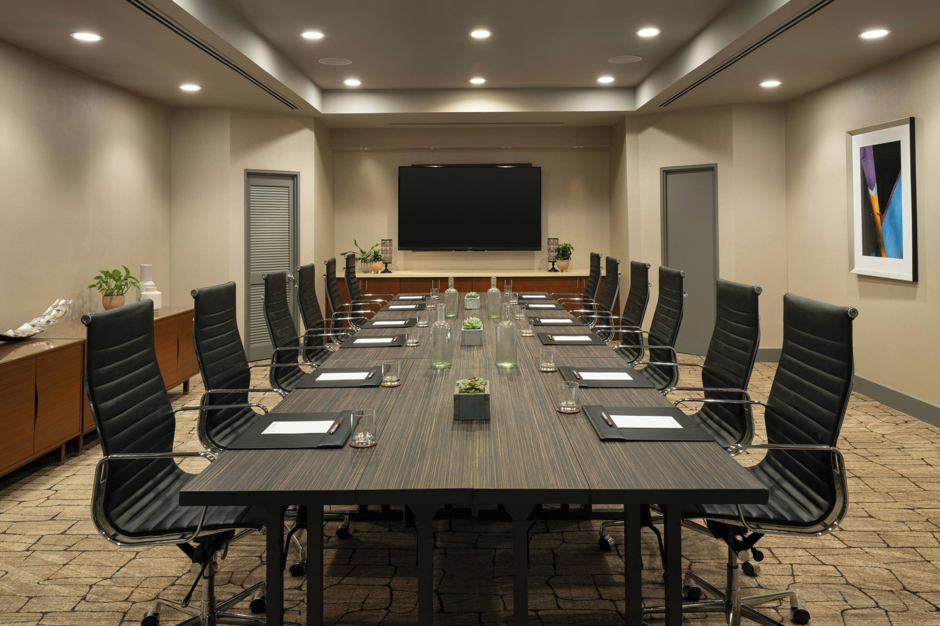 A conference room with a long table, black chairs, and a large screen.