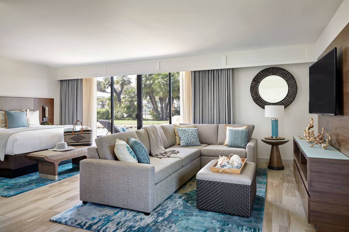 A modern hotel room with a large sectional sofa, a bed, a TV, and a view of palm trees.