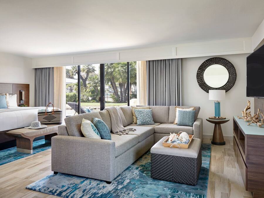 A modern hotel room with a large sectional sofa, a bed, a TV, and a view of palm trees.