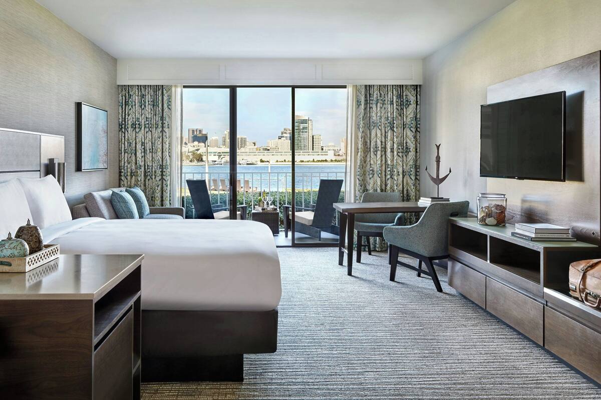 A modern hotel room with a large bed, a desk, a chair, a TV, and a view of the city skyline.