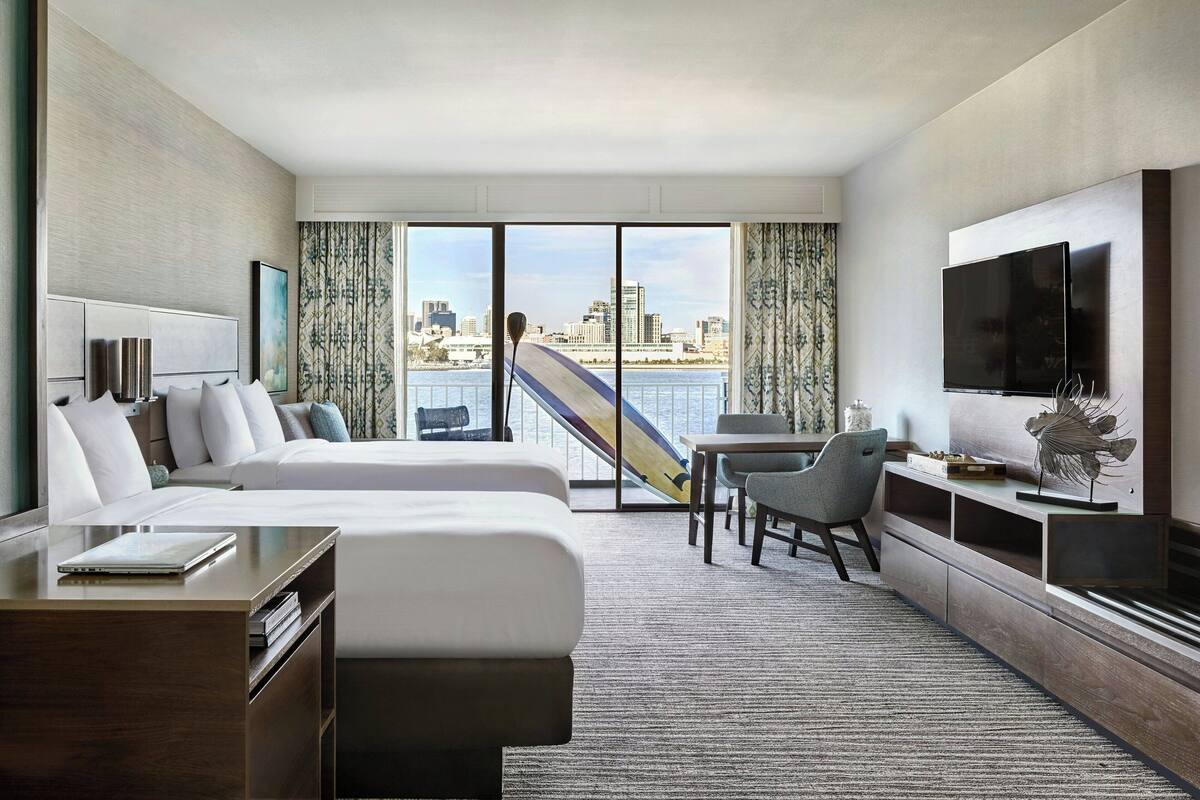 A hotel room with a bed, a desk, a chair, a TV, and a view of a cityscape through large windows.