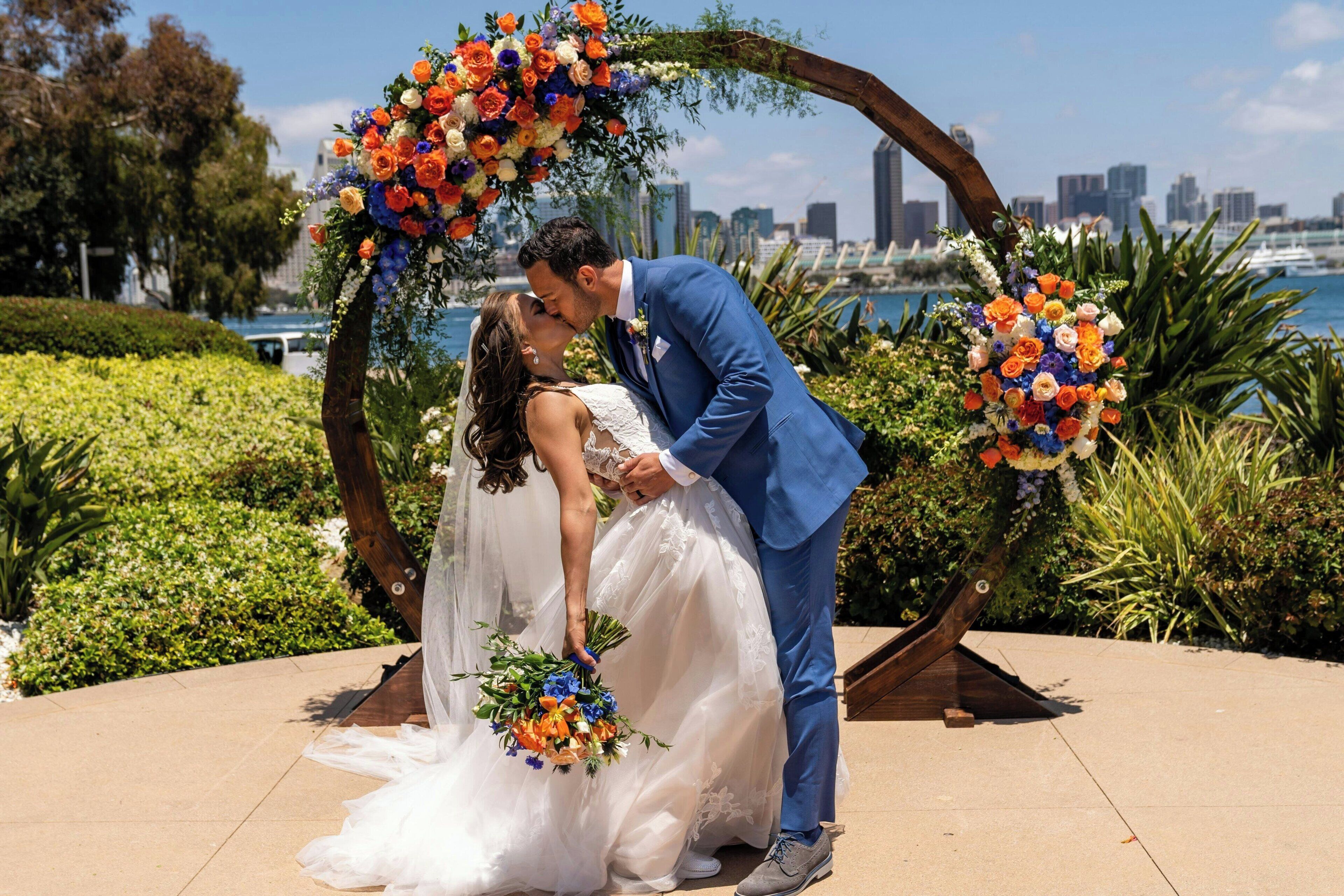 A couple kissing under a floral arch with a city skyline in the background.
