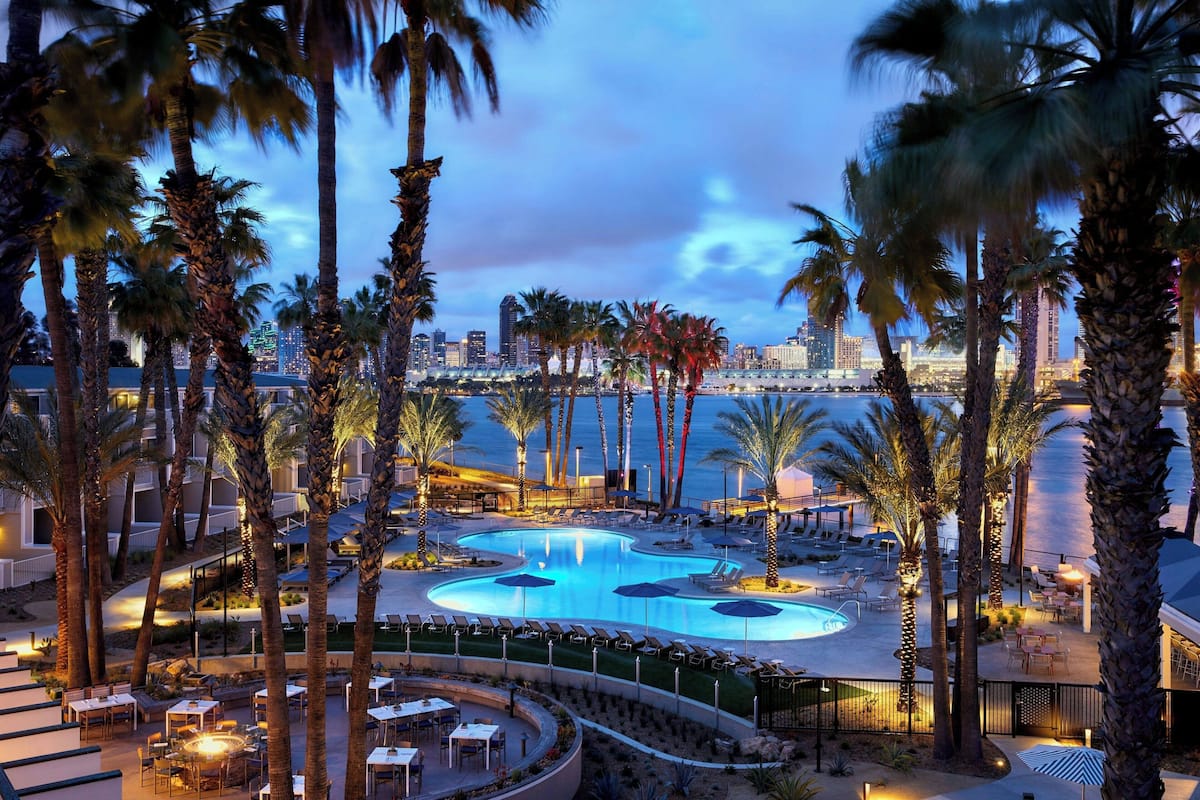 A resort pool area with palm trees, lounge chairs, and a city skyline in the background.
