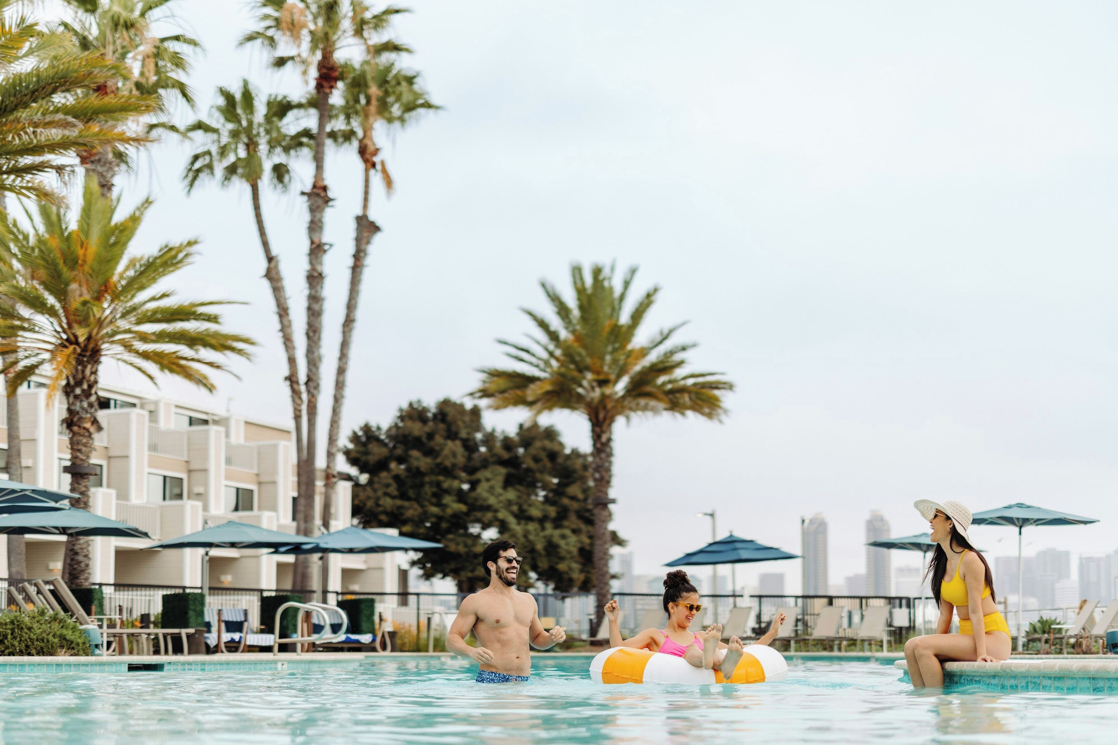 Una piscina con palmeras y sombrillas, rodeada de edificios.