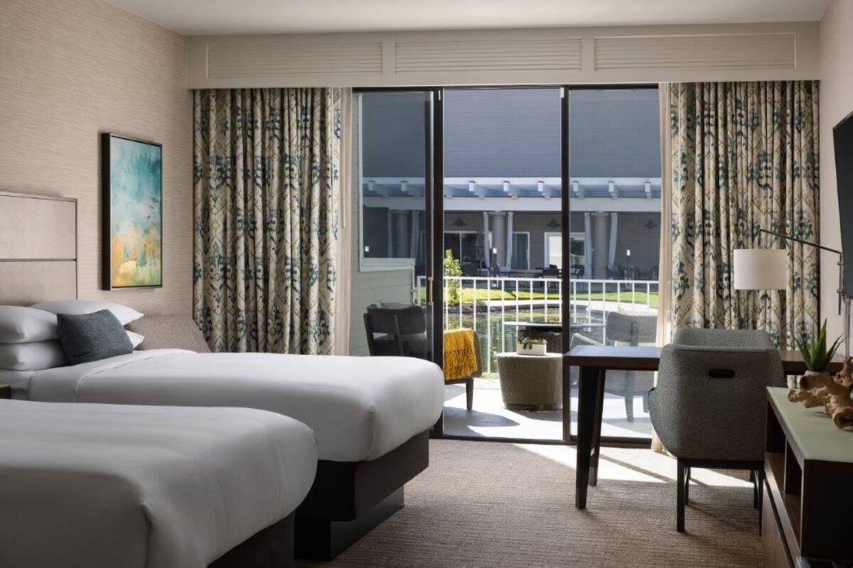 A hotel room with two beds, a desk, a chair, and a view of a building and a pool.