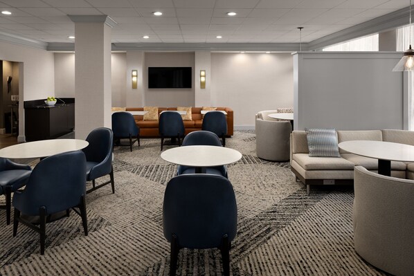A modern hotel lobby with a seating area, a flat-screen TV mounted on the wall, and a small kitchen counter with a bowl of fruit.