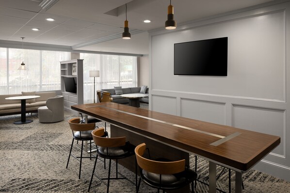 Un salón de conferencias moderno con una larga mesa de madera, cuatro taburetes altos, un sofá y un televisor montado en la pared.