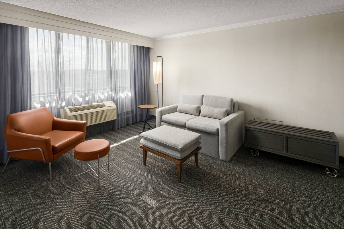 Habitación de hotel con sofá, sillón, reposapiés, lámpara de pie, radiador y ventana grande con cortinas.