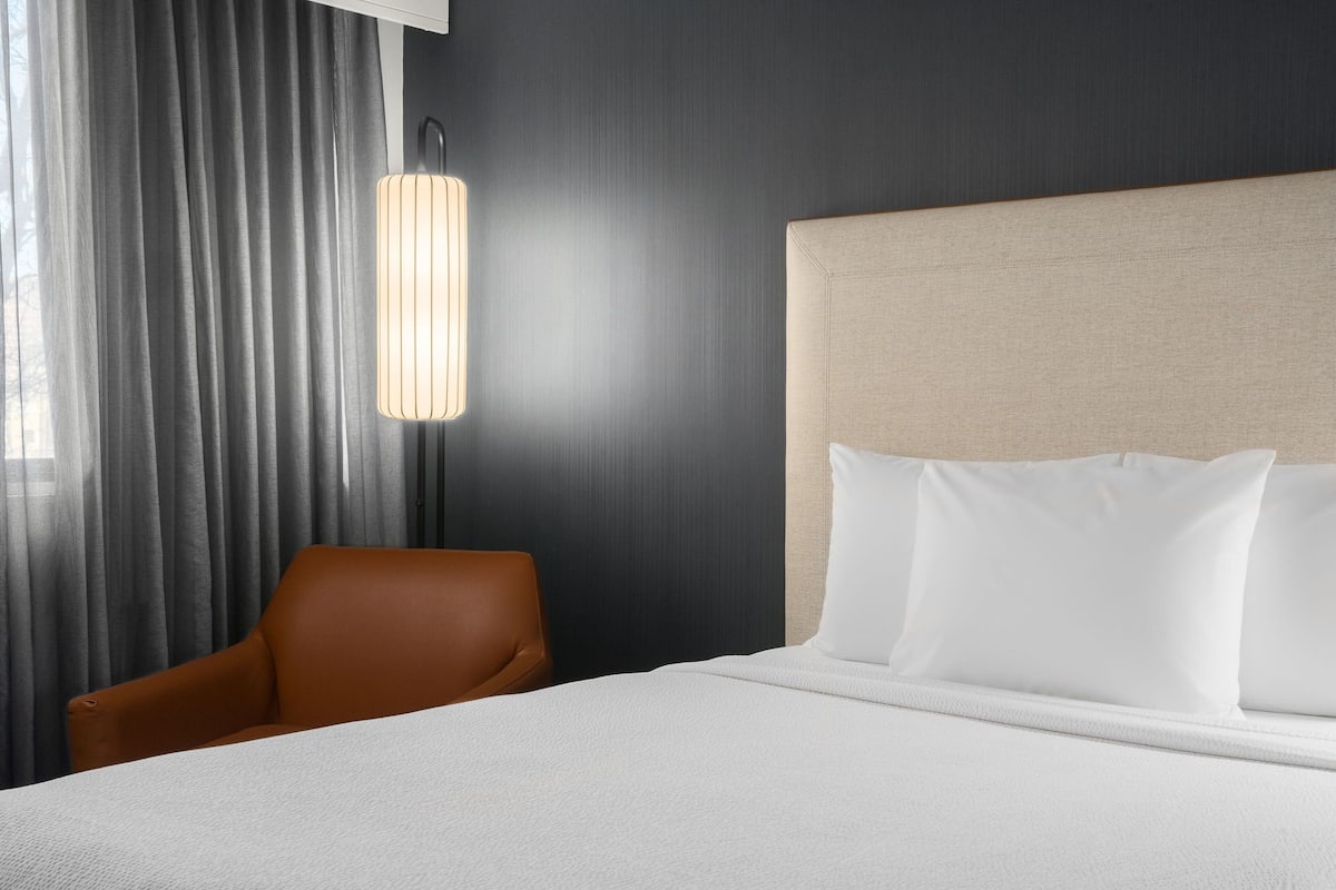 Habitación de hotel con una cama, una silla, una lámpara y vistas al exterior a través de cortinas.