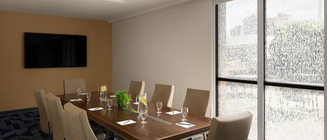A conference room with a long table, six chairs, a TV on the wall, and large windows with a view of trees.