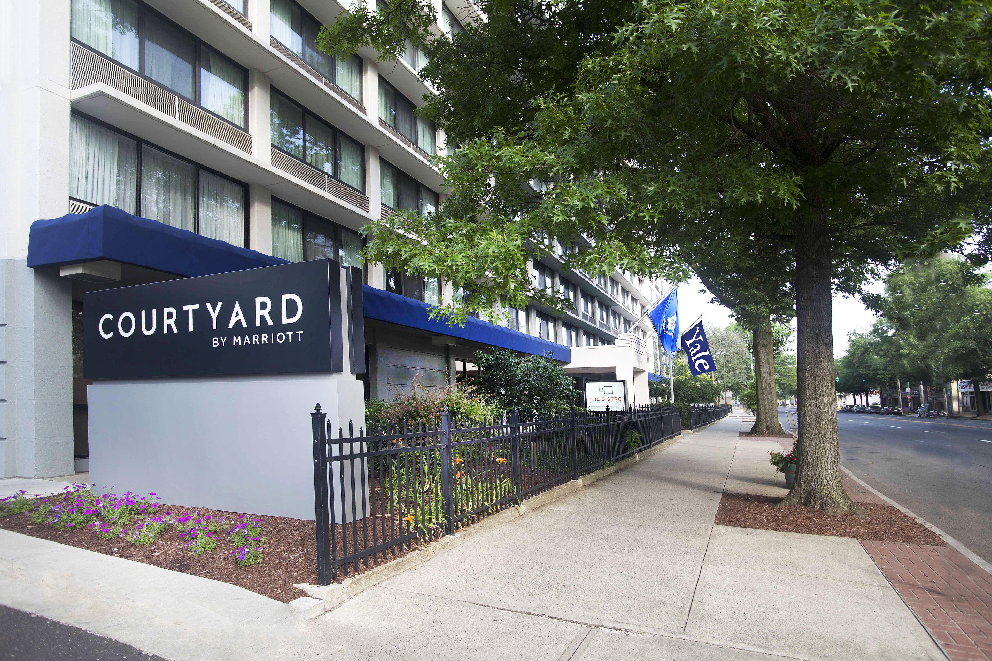 A Courtyard by Marriott hotel facade with a blue awning, a black metal fence, and a tree-lined sidewalk.