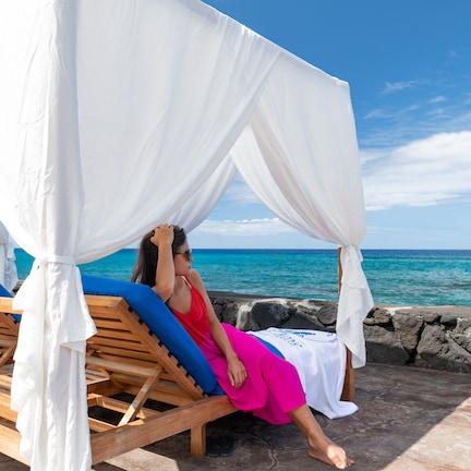 A person relaxing in a wooden lounge chair with a white canopy, by a stone wall overlooking the ocean.
