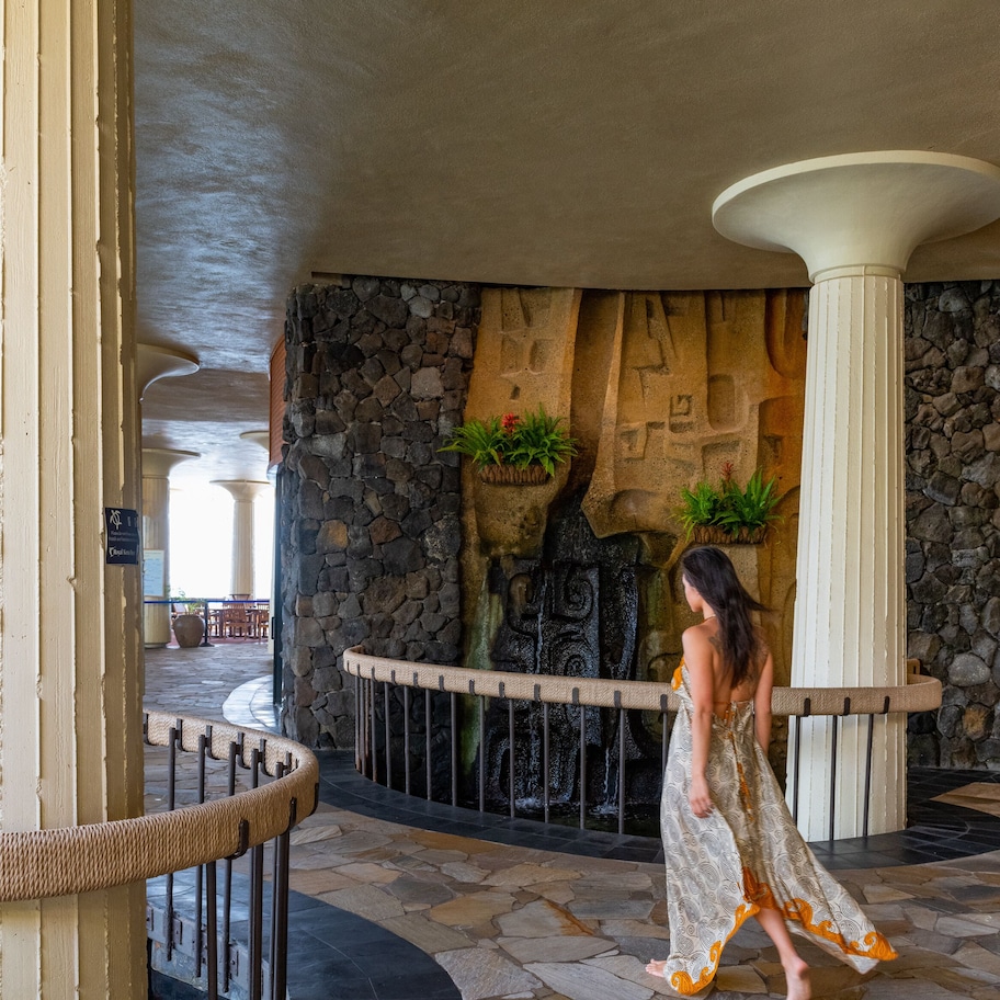 A woman walking in a lobby with stone walls, a fountain, and a seating area in the background.