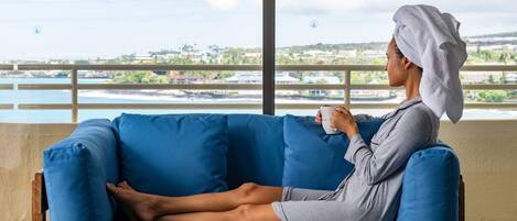 A person sitting on a blue sofa with a towel on their head, holding a cup, in a room with a large window overlooking a body of water and buildings.