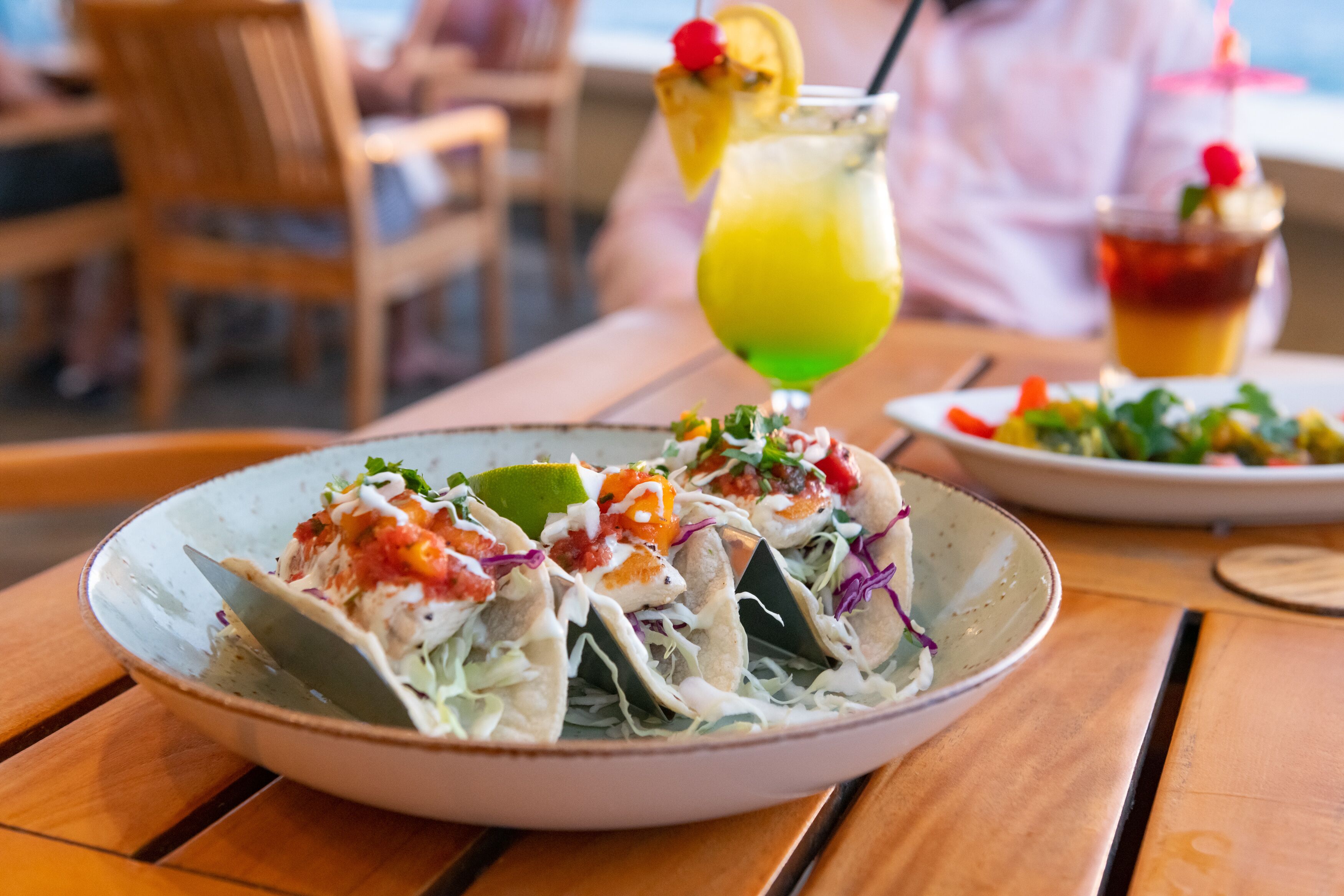 A plate of tacos with a lime wedge on the side, a green cocktail with a straw, and a glass of iced tea with a cherry on the rim.