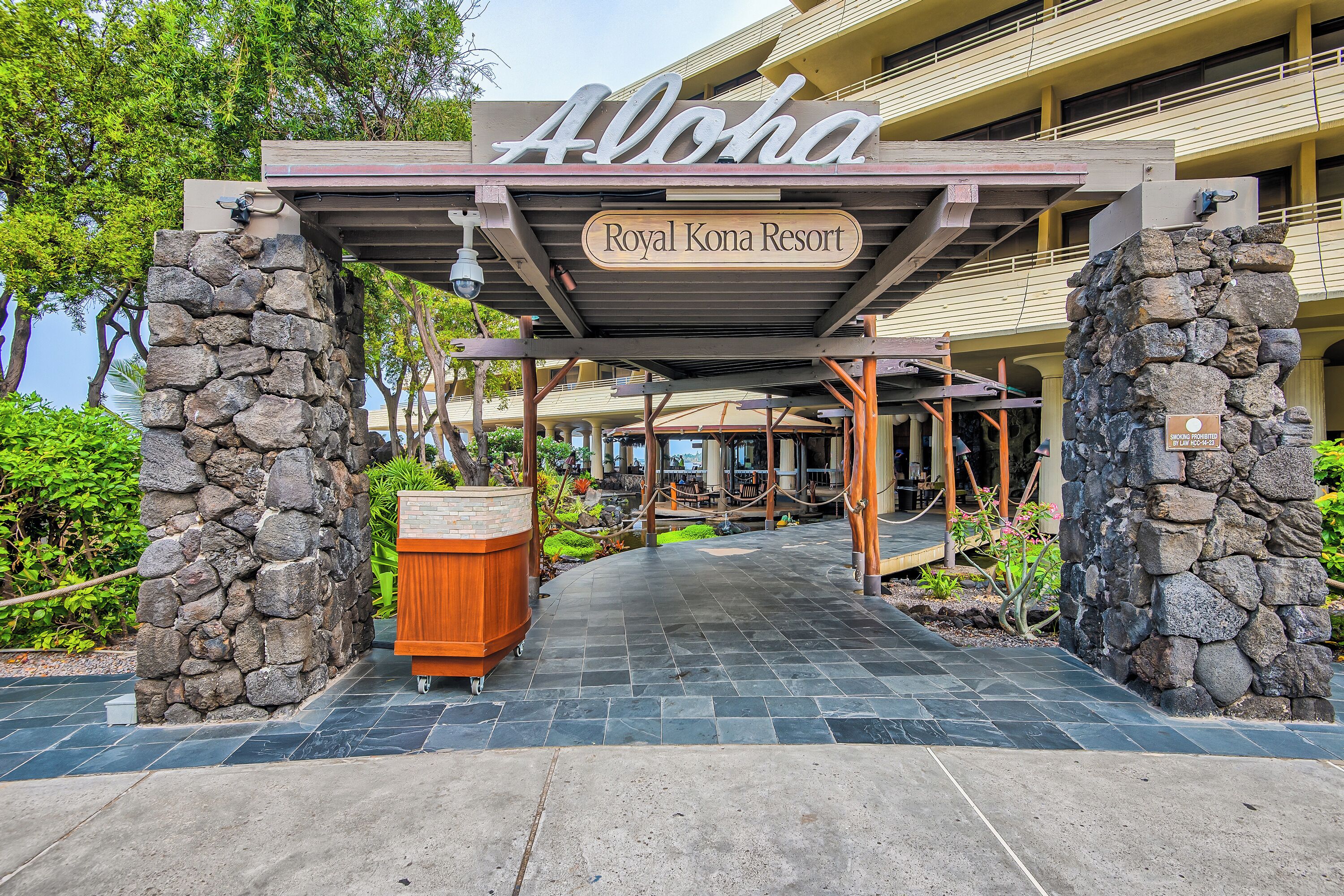 Entrada de un hotel con un letrero que dice 'Aloha Royal Kona Resort', pilares de piedra, un corredor cubierto y una planta en maceta.