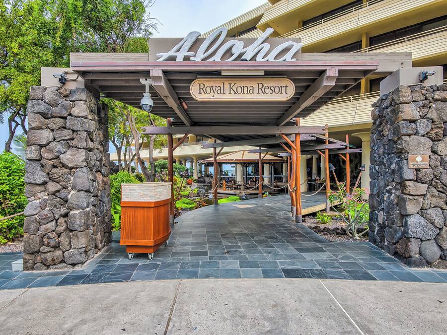 Entrada de un hotel con un letrero que dice 'Aloha Royal Kona Resort', pilares de piedra, un corredor cubierto y una planta en maceta.