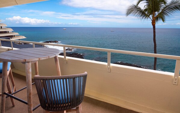 A balcony with a wooden table and chair, overlooking the ocean with boats and a palm tree.