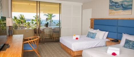 Ein Hotelzimmer mit zwei Betten, einem Schreibtisch und einem Balkon mit Blick auf Palmen und das Meer.