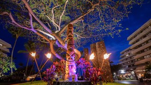 A night scene with a large tree, a tiki torch sculpture, and a stone structure. - Royal Kona Resort (Kailua-Kona)