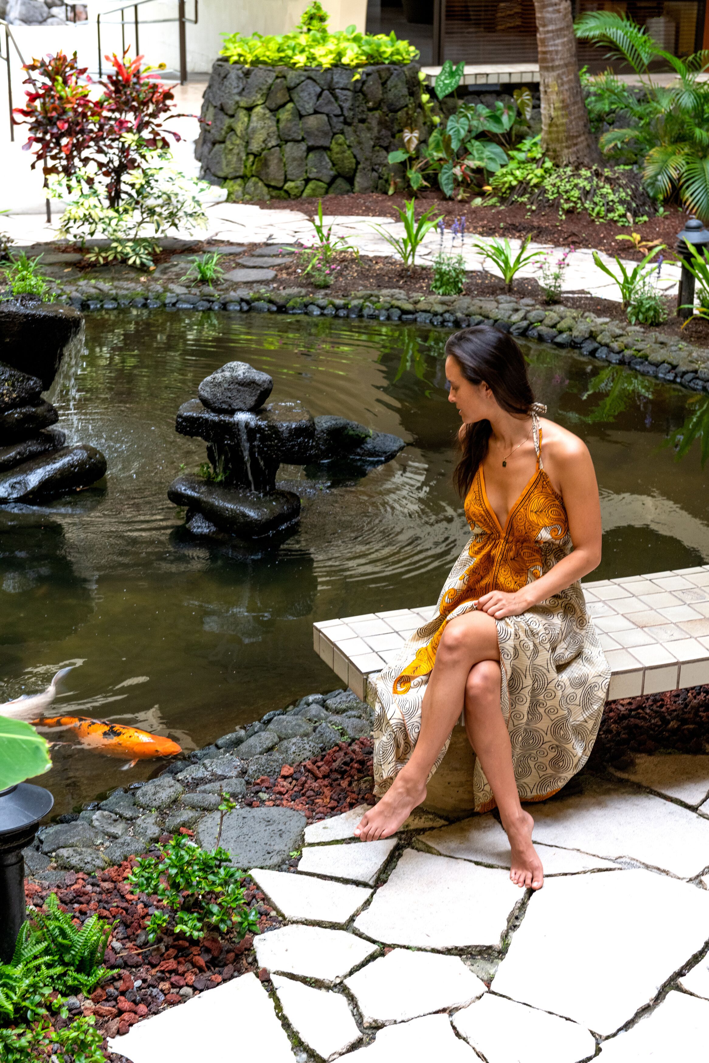 Una mujer sentada junto a un estanque con peces koi, rodeada de un jardín con diversas plantas y un muro de piedra.