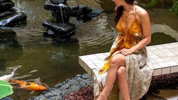 Una mujer sentada junto a un estanque con peces koi, rodeada de un jardín con diversas plantas y un muro de piedra.