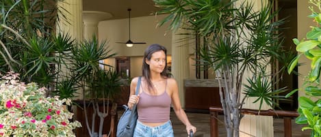 A woman walking with a suitcase in a hotel lobby with stone flooring, columns, and potted plants.