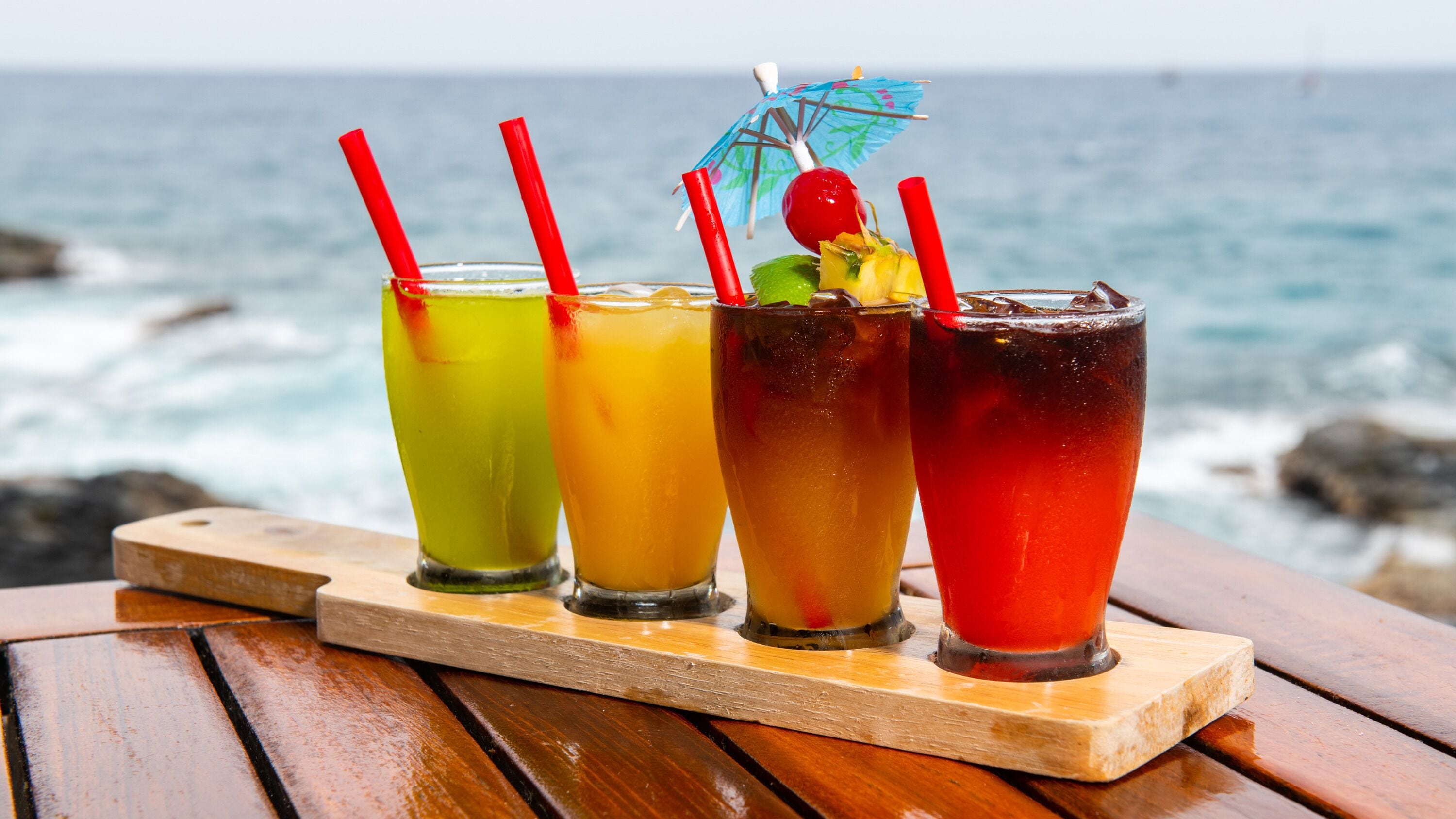 Four colorful drinks on a wooden tray by the ocean.