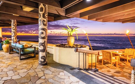 An indoor area with a view of the ocean, stone flooring, wooden furniture, and decorative columns.