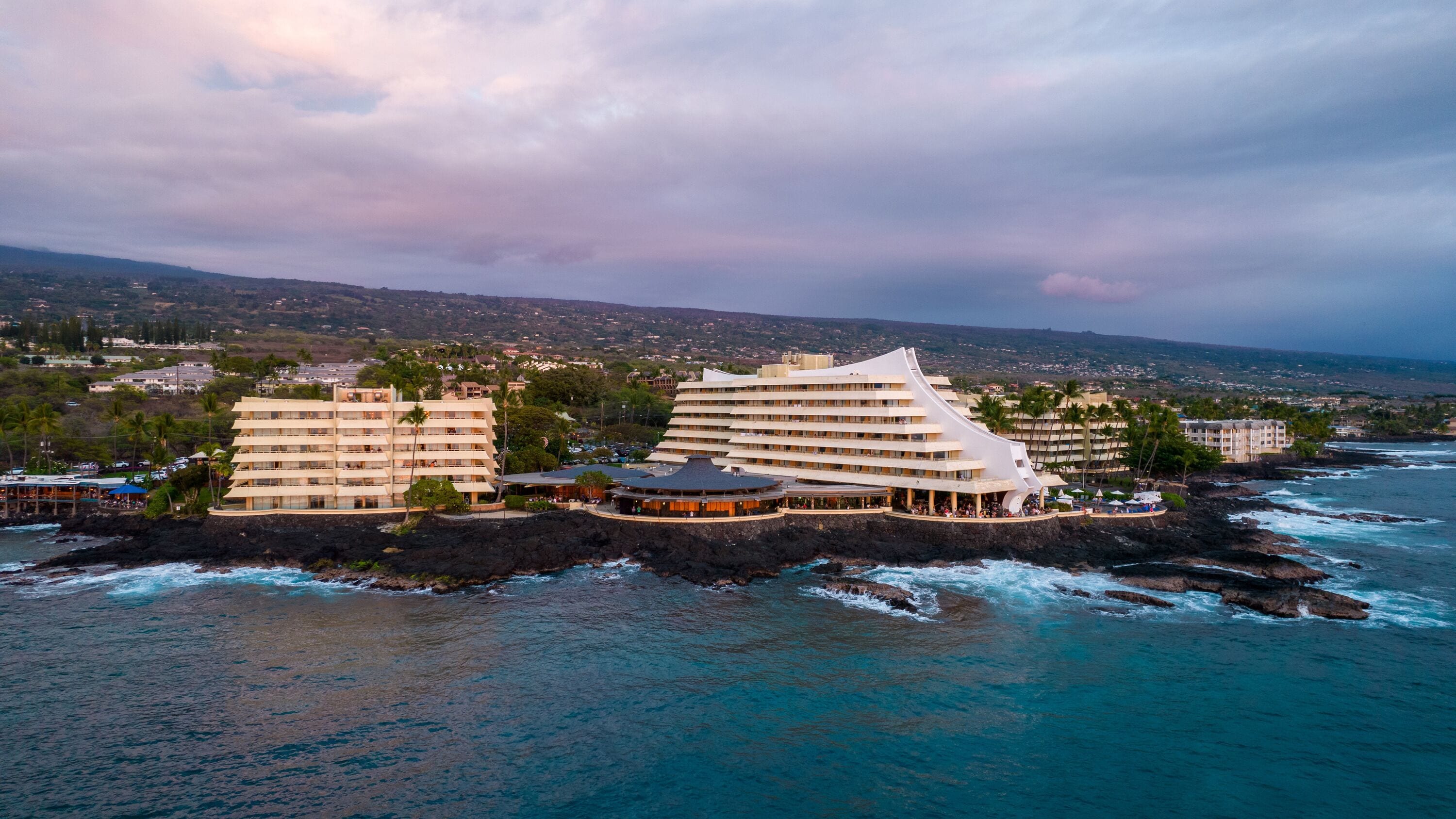 A coastal hotel with multiple buildings, a beachfront, and a view of the ocean and distant hills.