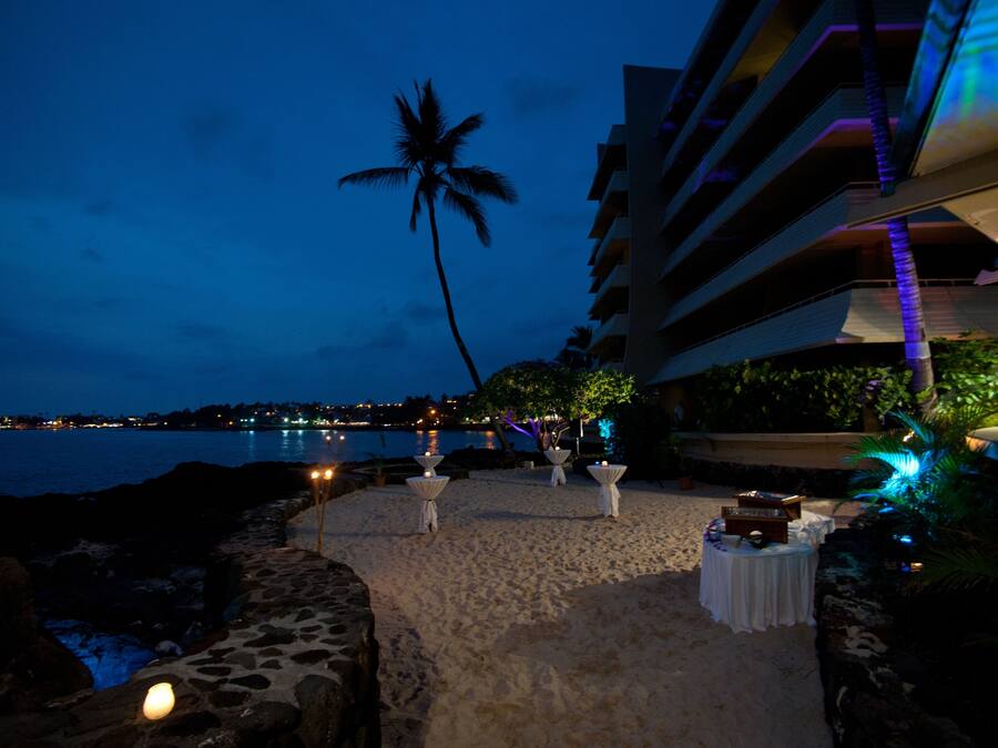 A beachside view with a palm tree, a stone pathway, and tables with white cloths.