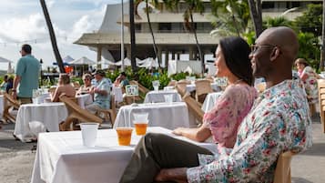 A beachside dining area with tables and chairs, palm trees, and a building in the background.