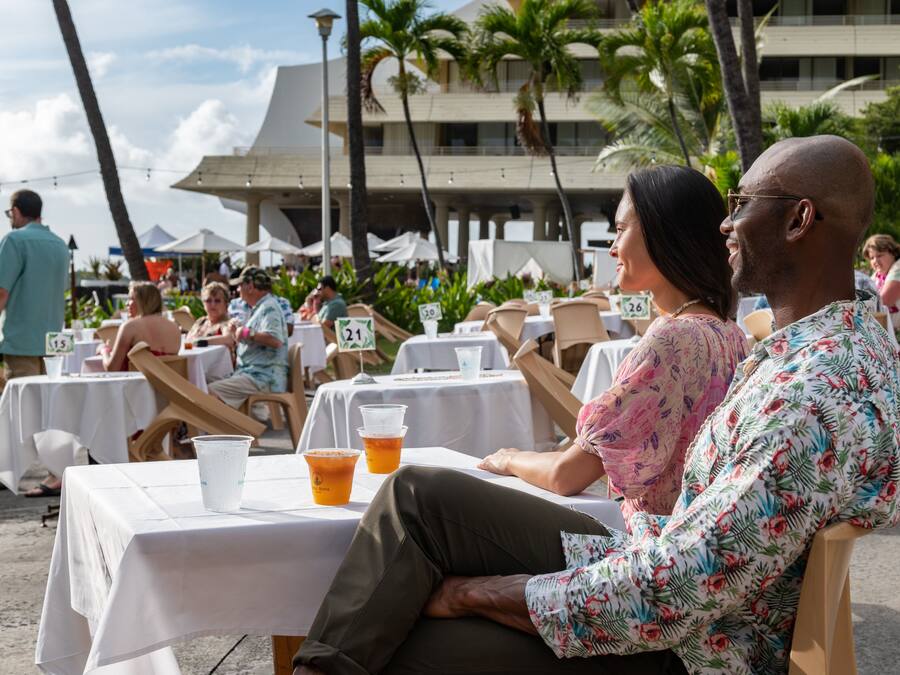 A beachside dining area with tables and chairs, palm trees, and a building in the background.