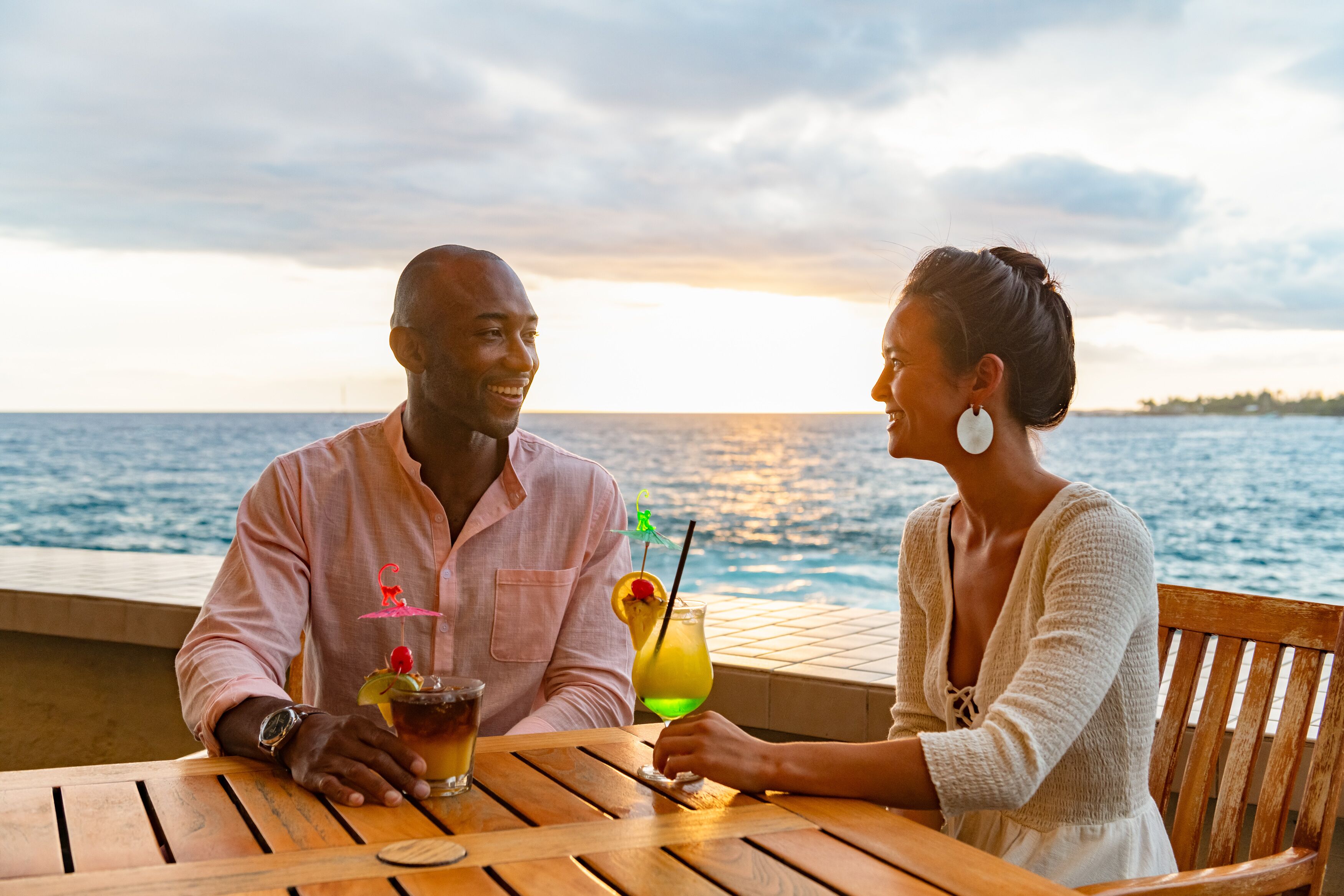 A couple seated at a wooden table by a waterfront, with drinks and a sunset view.